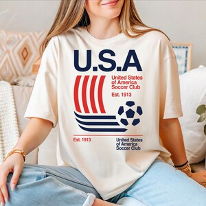 A woman wearing a white t-shirt with the text "U.S.A" and a soccer ball graphic, sitting on a couch.