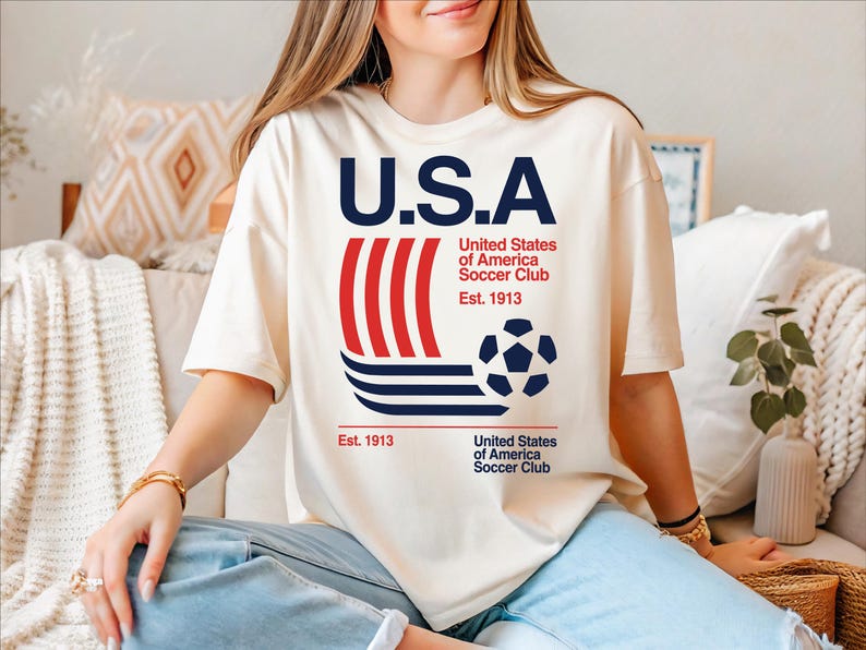 A woman wearing a white t-shirt with the text "U.S.A" and a soccer ball graphic, sitting on a couch.