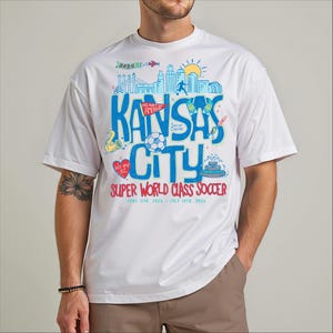 A white t-shirt with a colorful graphic design featuring various elements related to Kansas City, including a city skyline, a soccer ball, and other sports-related imagery.
