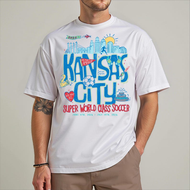 A white t-shirt with a colorful graphic design featuring various elements related to Kansas City, including a city skyline, a soccer ball, and other sports-related imagery.