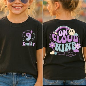 May include: Black t-shirt with a retro design. The front features the number 9, the name "Emily", and star accents. The back displays "On Cloud Nine" with a rainbow, smiley face, flowers, and clouds. A fun, colorful tee.