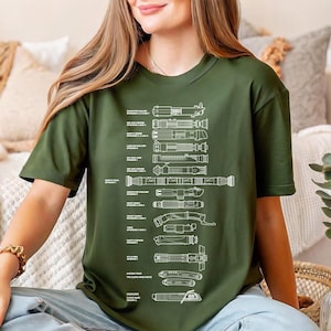 May include: Olive green t-shirt featuring a detailed white diagram of various lightsaber designs. The diagram includes text labels for each lightsaber. The shirt is a casual, short-sleeved style.