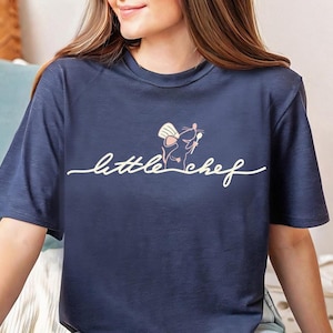 May include: Dark blue t-shirt with a white cursive "little chef" design. The graphic shows a mouse in a chef's hat holding utensils. The shirt has a crew neck and short sleeves.