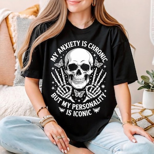 Puede incluir: Camiseta negra con un diseño de calavera, mariposas y el texto "MY ANXIETY IS CHRONIC BUT MY PERSONALITY IS ICONIC". La camiseta es de manga corta y cuello redondo.