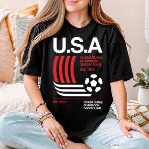 A woman wearing a black t-shirt with a graphic design of a soccer ball and stripes, sitting on a couch with a potted plant in the background.