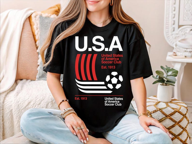 A woman wearing a black t-shirt with a graphic design of a soccer ball and stripes, sitting on a couch with a potted plant in the background.