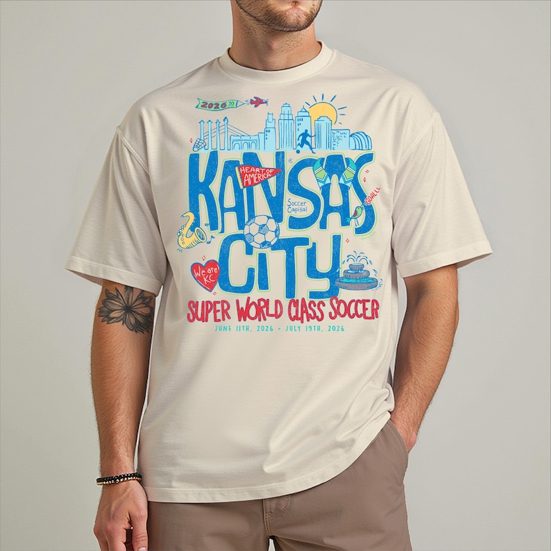 A man wearing a white t-shirt with a graphic design featuring various elements related to Kansas City, including a city skyline, a soccer ball, and other symbols.