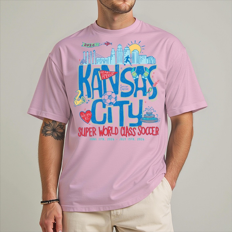 A man wearing a pink t-shirt with a graphic design featuring various elements related to Kansas City, including a city skyline, a soccer ball, and other symbols.