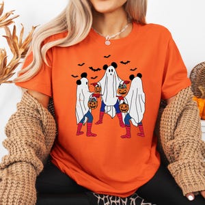 A woman wearing an orange t-shirt with a Halloween-themed graphic of three ghostly figures holding pumpkins and wearing spider-like legs.