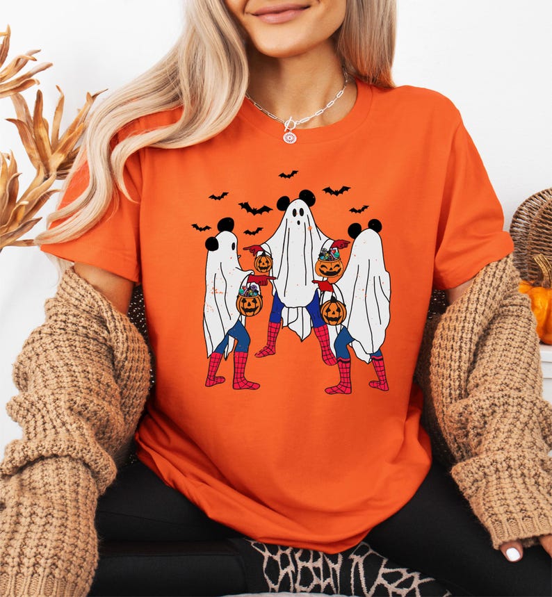 A woman wearing an orange t-shirt with a Halloween-themed graphic of three ghostly figures holding pumpkins and wearing spider-like legs.