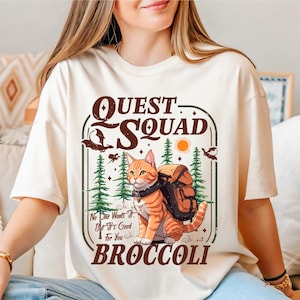 Może przedstawiać: Kremowa koszulka z grafiką przedstawiającą kreskówkowego kota z plecakiem, na tle leśnej scenerii. Tekst brzmi "Quest Squad" i "Broccoli". Dodatkowy tekst mówi "No One Wants It, But It's Good For You".
