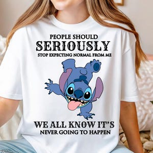 May include: White t-shirt featuring a blue cartoon character with large eyes and a pink tongue. The shirt has the text "PEOPLE SHOULD SERIOUSLY STOP EXPECTING NORMAL FROM ME WE ALL KNOW IT'S NEVER GOING TO HAPPEN" in black.