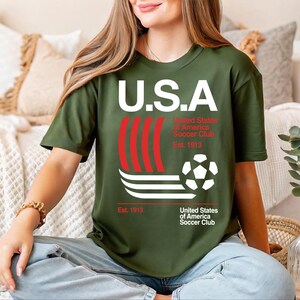 A woman wearing a green t-shirt with the text "U.S.A. United States of America Soccer Club Est. 1913" is sitting on a couch.