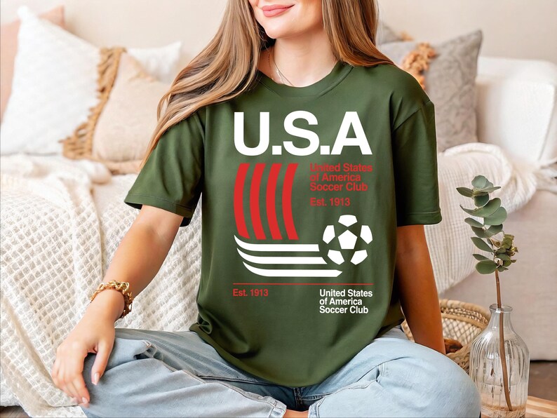 A woman wearing a green t-shirt with the text "U.S.A. United States of America Soccer Club Est. 1913" is sitting on a couch.