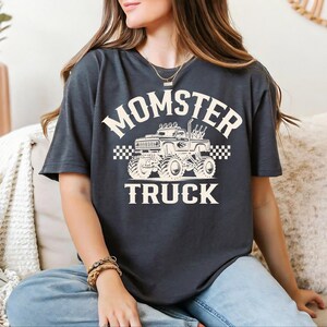 A woman wearing a black t-shirt with a graphic of a monster truck and the text "MOMSTER TRUCK" is sitting on a couch.