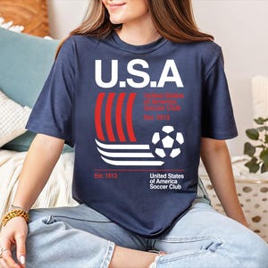 A woman wearing a blue t-shirt with a graphic design of a soccer ball and stripes, sitting on a couch.