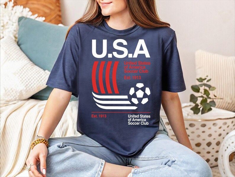 A woman wearing a blue t-shirt with a graphic design of a soccer ball and stripes, sitting on a couch.