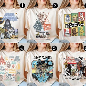 May include: Cream-colored Star Wars graphic t-shirts. Each shirt features a different design with characters and logos from the Star Wars franchise. Designs include Darth Vader, Stormtroopers, and the phrase "May the 4th be with you."