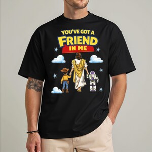 A black t-shirt with a graphic design featuring a cartoon character holding hands with two other characters, one of whom is Jesus. The text on the t-shirt reads "You've Got a Friend in Me".
