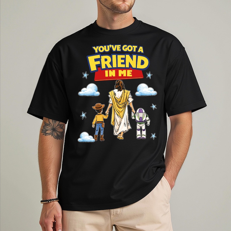 A black t-shirt with a graphic design featuring a cartoon character holding hands with two other characters, one of whom is Jesus. The text on the t-shirt reads "You've Got a Friend in Me".