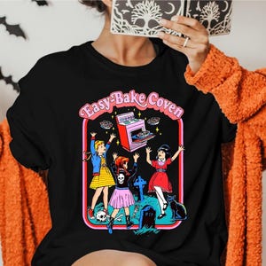 May include: Black t-shirt with a colorful graphic featuring the text "Easy Bake Coven" in pink. The graphic depicts three cartoon figures with a retro oven, a tombstone, and a black cat. The shirt is worn by a person with an orange sweater.