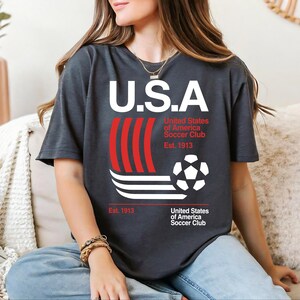 A woman wearing a black t-shirt with a graphic design of a soccer ball and stripes, sitting on a couch.