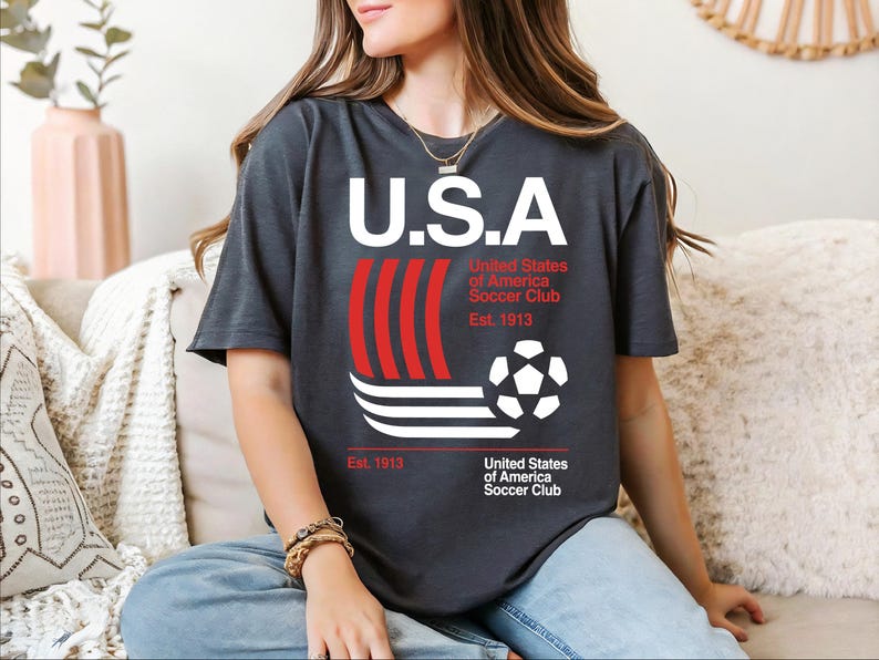 A woman wearing a black t-shirt with a graphic design of a soccer ball and stripes, sitting on a couch.