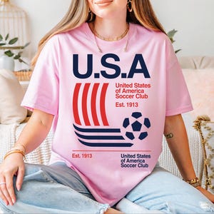 A woman wearing a pink t-shirt with the text "U.S.A United States of America Soccer Club Est. 1913" printed on it, sitting on a couch.