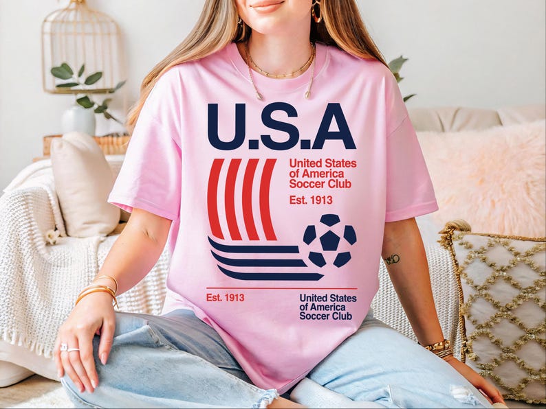 A woman wearing a pink t-shirt with the text "U.S.A United States of America Soccer Club Est. 1913" printed on it, sitting on a couch.