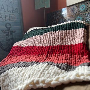 May include: A chunky knit blanket in a color block design. The blanket features stripes of white, gray, coral, red, pink, and dark green. The blanket is draped over a brown couch.