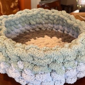 May include: A round, hand-knitted pet bed in shades of grey, green, and beige. The bed is made of a thick, plush yarn, creating a soft and cosy texture. The design features a layered effect with a textured, raised edge.