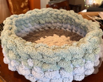 May include: A round, hand-knitted pet bed in shades of grey, green, and beige. The bed is made of a thick, plush yarn, creating a soft and cosy texture. The design features a layered effect with a textured, raised edge.