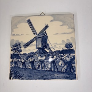May include: Square ceramic tile featuring a blue and white illustration of a windmill in a rural landscape. The artwork depicts a windmill with large blades, haystacks, and a cloudy sky. The tile has a metal hanger.