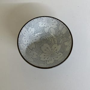 以下が含まれることがあります： 黒い縁取りと灰色の花柄が施された白いセラミックボウル