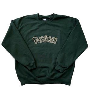 Könnte beinhalten: Dunkelgrünes Sweatshirt mit dem Wort "Pokémon" in goldfarbener, stilisierter Schrift auf der Brust. Das Sweatshirt hat einen Rundhalsausschnitt und lange Ärmel. Das Kleidungsstück wird auf weißem Hintergrund präsentiert.