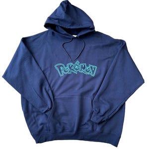 Könnte beinhalten: Marineblauer Kapuzenpullover mit Fronttasche und Kordelzugkapuze. Das Wort "Pokémon" ist in einer türkisfarbenen, umrandeten Schrift auf die Brust gedruckt. Der Pullover ist aus weichem Material.