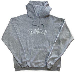 Könnte beinhalten: Grauer Kapuzenpullover mit dem Wort "Pokémon" in Weiß auf der Brust. Der Pullover hat eine Vordertasche und weißen Text am linken Ärmel. Der Hoodie ist aus einem weichen Material.
