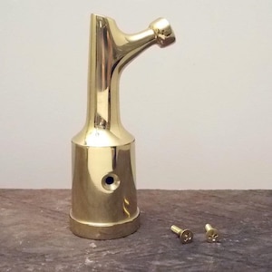 May include: A shiny, gold-colored metal bracket with a curved arm and a rounded end. Two small screws are next to the bracket. The bracket has a cylindrical base and a small blue dot. The item is likely for home decor or hardware.