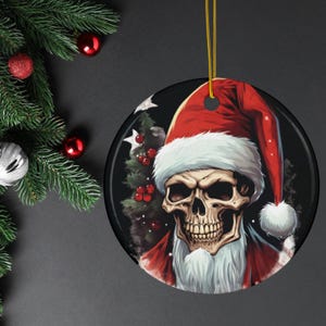 Puede incluir: Adorno navideño redondo con una calavera que lleva un gorro y barba de Papá Noel. El adorno tiene un fondo negro con un árbol de Navidad y bayas rojas. Una cuerda dorada está unida para colgarlo.