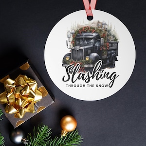 Könnte beinhalten: Ein weißer Weihnachtsschmuck mit einer Illustration eines schwarzen Trucks, verziert mit einem Kranz und Lichtern, und dem Text "Slashing Through The Snow". Der Schmuck hängt an einem roten Band. Eine Geschenkbox mit goldener Schleife und Weihnachtsschmuck befindet sich im Hintergrund.