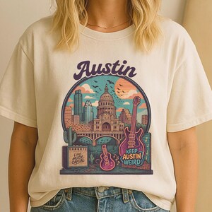 Puede incluir: Camiseta color crema con un gráfico colorido de Austin, Texas. El diseño incluye el horizonte de la ciudad, una guitarra y el texto "Keep Austin Weird". La camiseta también tiene el texto "Live Music Capital."