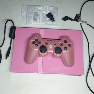 May include: A pink Sony PlayStation 2 console with a matching metallic pink controller, a black power brick with a cord, a separate black power cable with a plug, and a small clear plastic bag containing a black adapter component.