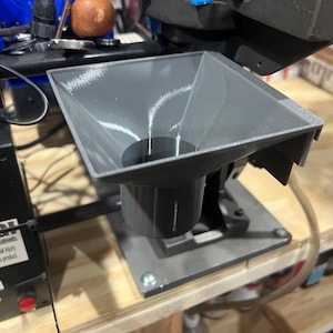 May include: A gray plastic funnel with a square top and a cylindrical base is attached to a metal machine. The funnel is designed to direct materials into the machine. The image is taken from a close-up angle.