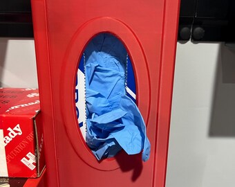 May include: A red plastic glove dispenser mounted on a wall. The dispenser has an oval opening revealing blue gloves inside. The dispenser is rectangular with a curved top. Other boxes and items are visible in the background.