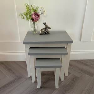 May include: A set of three grey nesting tables with white legs. The tables are made of wood and have a distressed finish. The top table has a small decorative rabbit figurine on it.