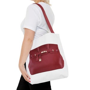May include: White tote bag featuring a large image of a burgundy handbag with gold-colored hardware. The bag has a shoulder strap in a matching burgundy color. The tote bag is carried over the shoulder.