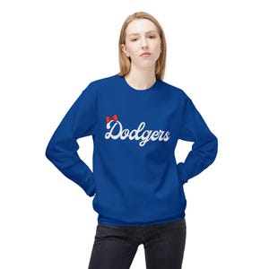 Peut inclure: Un sweat-shirt bleu roi avec le mot "Dodgers" en écriture blanche. Un petit nœud rouge est au-dessus du mot. Le sweat-shirt est de style col rond.