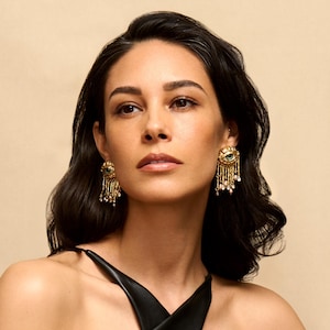 May include: A woman with long dark hair wearing a black sleeveless top and gold earrings with green and blue eye designs.