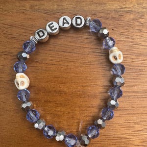 Pode incluir: Uma pulseira de contas com contas facetadas roxo-claras translúcidas, contas metálicas prateadas, duas pequenas contas em forma de caveira na cor branco sujo e quatro contas de letras prateadas que soletram a palavra DEAD em preto.