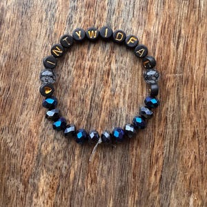 May include: A circular beaded bracelet featuring black letter beads with gold text spelling INTYWIDFA, interspersed with two grey marbled beads, a small gold heart bead, and a series of faceted dark blue and black beads along the bottom half.